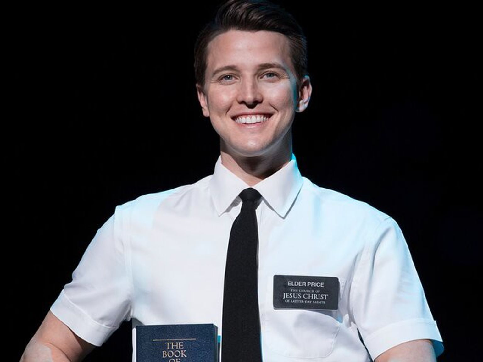 En leende ung man i vit skjorta och svart slips, som bär en namnbricka med texten "Elder Price" och håller i en bok med titeln "The Book of Mormon".