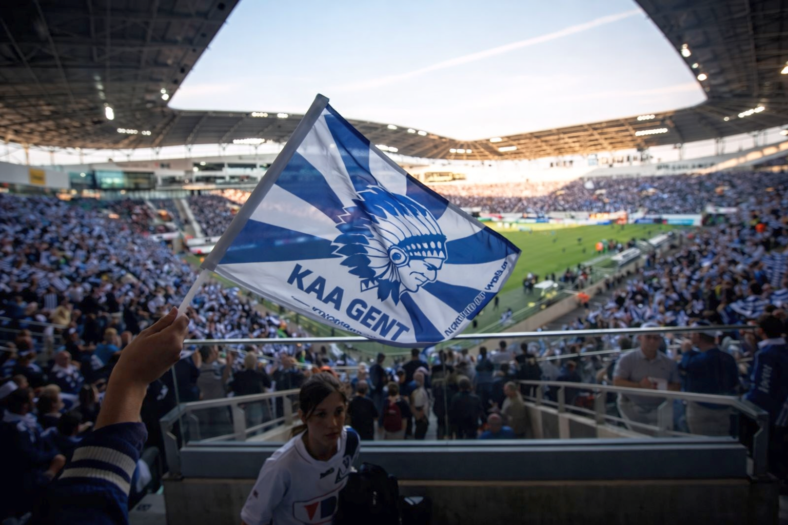 A KAA Gent flag with a Native American chief logo waves in a crowded football stadium.