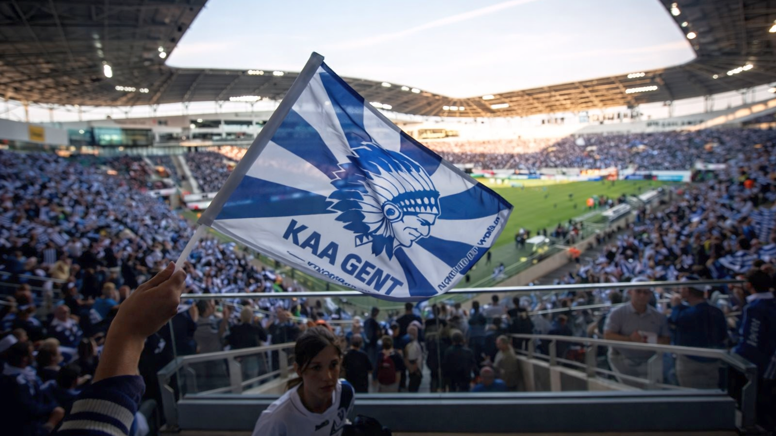 A KAA Gent flag with a Native American chief logo waves in a crowded football stadium.
