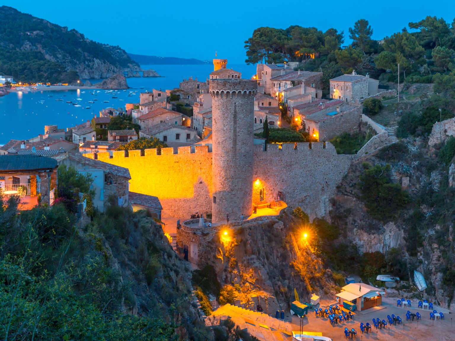 Vue nocturne d’une ancienne forteresse en pierre illuminée et d’une ville côtière surplombant une baie avec des bateaux. Tossa de Mar, Espagne
