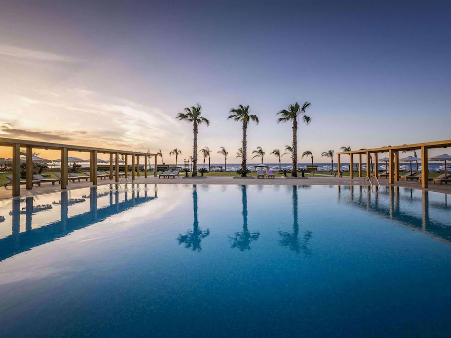 uma grande piscina rodeada por palmeiras e cadeiras ao pôr do sol, Mitsis Selection Alila