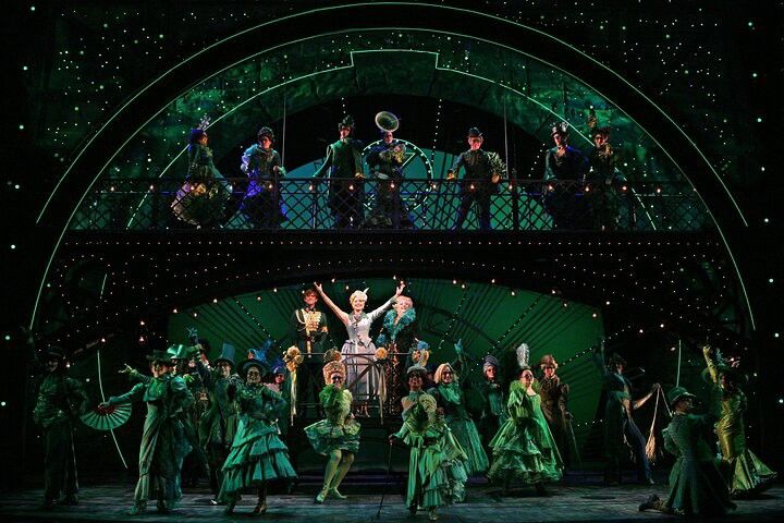 A musical stage with a large cast in elaborate green costumes on two levels, under green sparkling lights.
