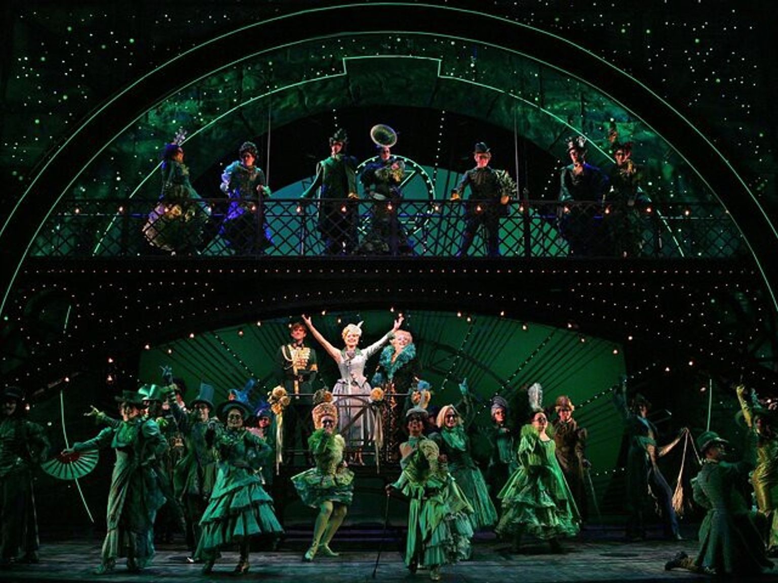 A musical stage with a large cast in elaborate green costumes on two levels, under green sparkling lights.