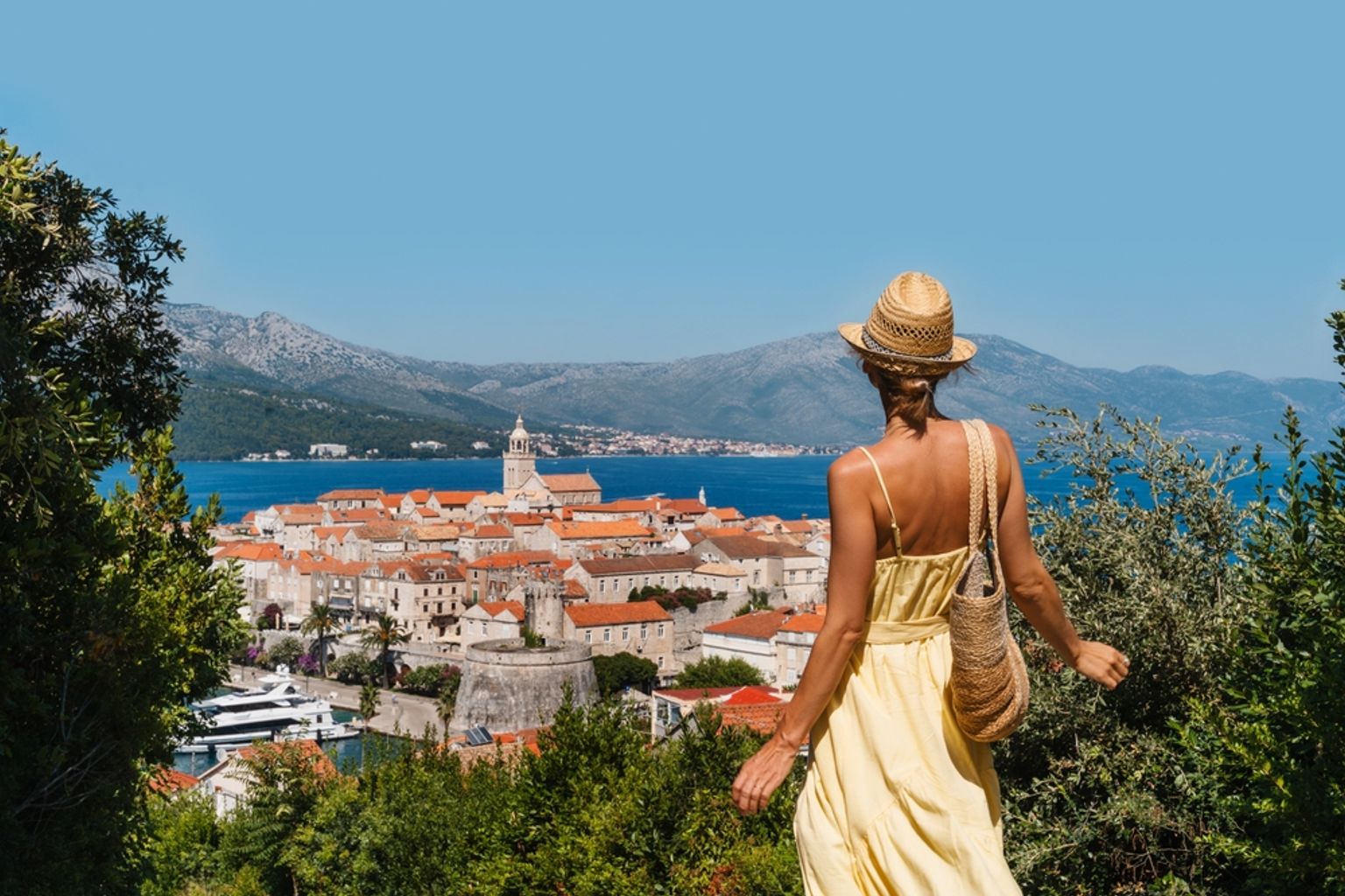 Woman in a yellow dress overlooks a coastal town with red roofs, a church, and a bay backed by mountains. Korčula, Croatia