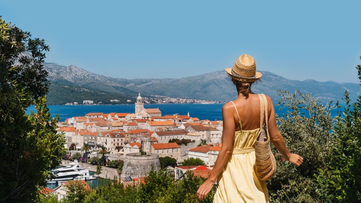 Woman in a yellow dress overlooks a coastal town with red roofs, a church, and a bay backed by mountains. Korčula, Croatia