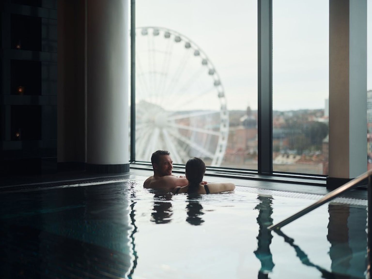 ein Mann und eine Frau sitzen in einem Schwimmbecken mit einem Riesenrad im Hintergrund.