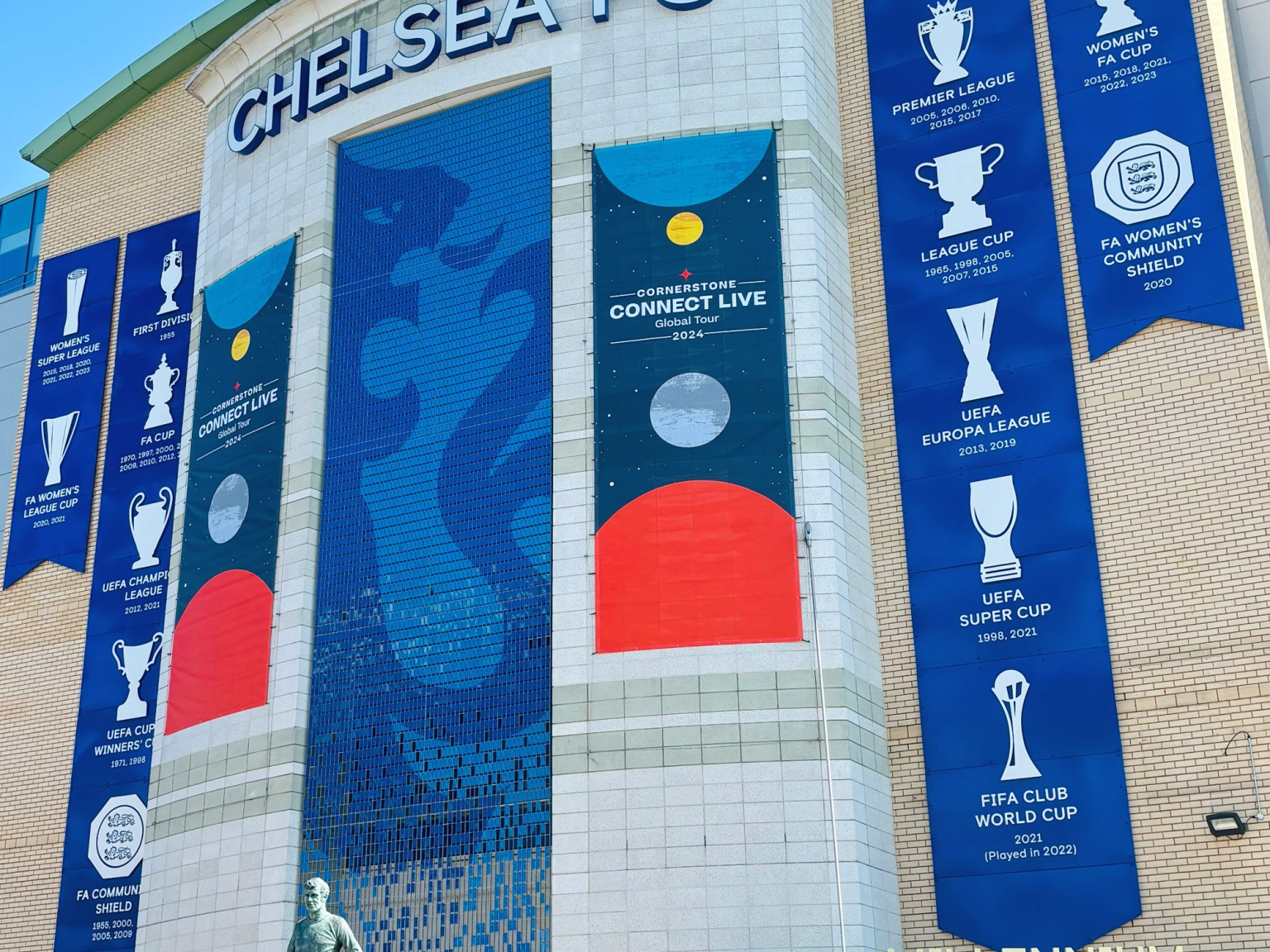 Utsiden av Chelsea FCs stadion, med klubbens logo, stort navn, bannere som viser ulike trofeer og en statue av en spiller.