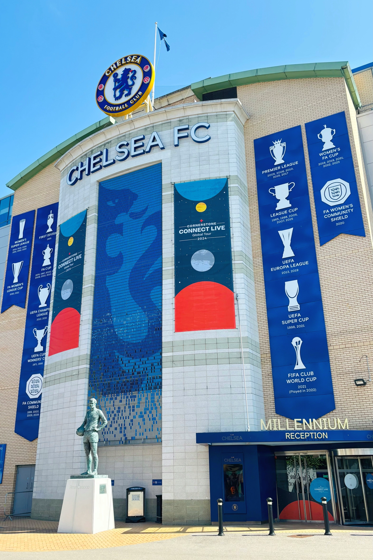 Utsiden av Chelsea FCs stadion, med klubbens logo, stort navn, bannere som viser ulike trofeer og en statue av en spiller.