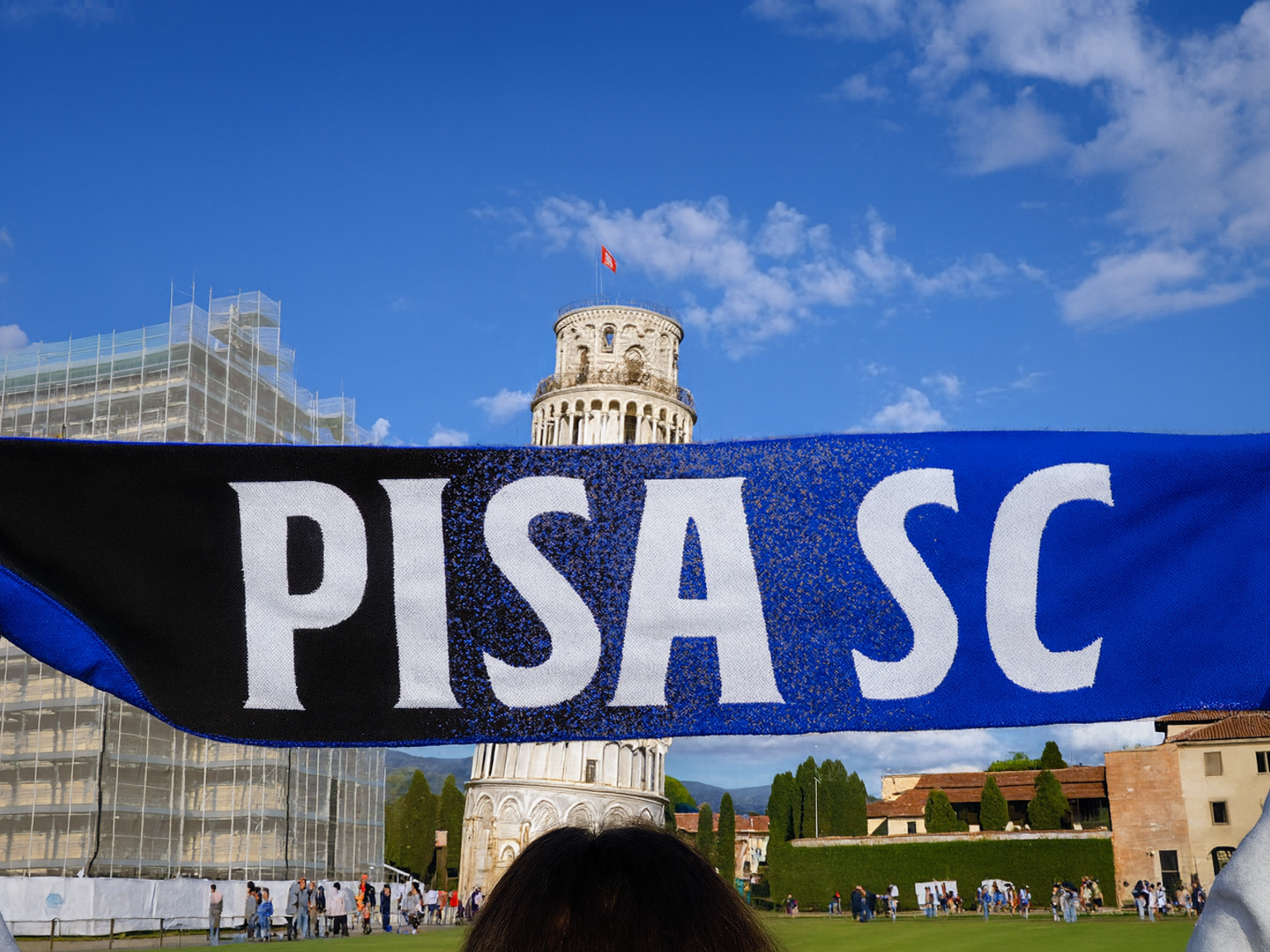 A fan holds a blue and black "PISA SC" scarf in front of the Leaning Tower of Pisa.