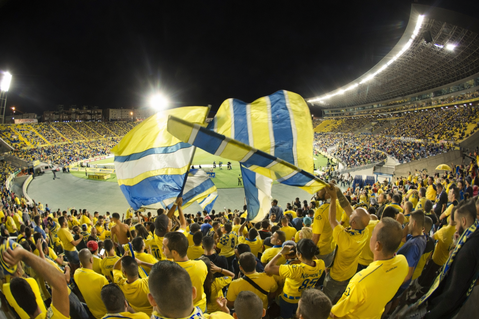 Un estadio por la noche, lleno de aficionados vestidos de amarillo ondeando grandes banderas amarillas y azules.
