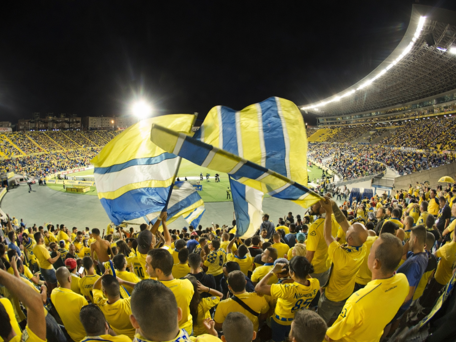 Un estadio por la noche, lleno de aficionados vestidos de amarillo ondeando grandes banderas amarillas y azules.