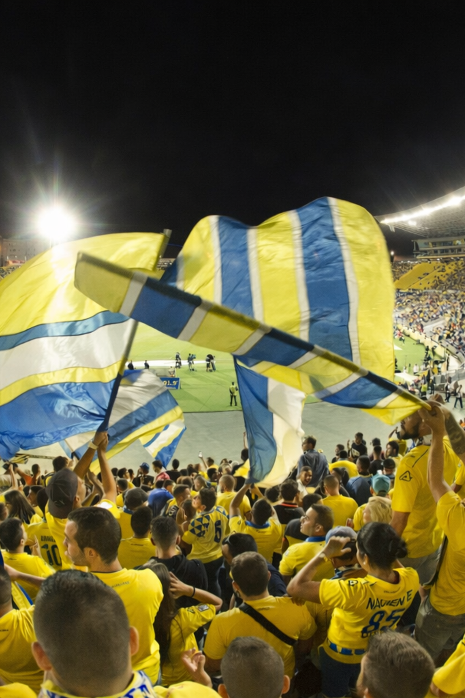 Un estadio por la noche, lleno de aficionados vestidos de amarillo ondeando grandes banderas amarillas y azules.