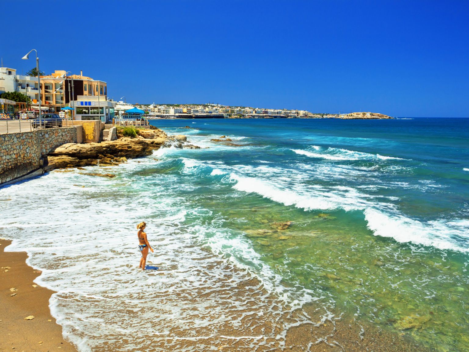En kvinde står i det lave, klart blå havvand på en solrig strand med bygninger og klipper langs kysten.