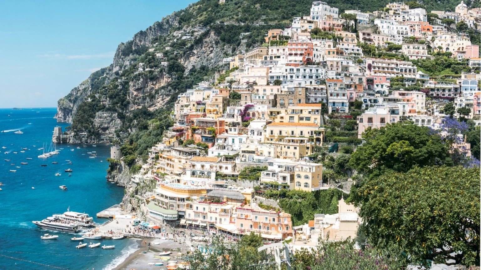Positano, Italy: vibrant, colorful houses tiered up a steep hillside overlooking a beach and blue sea with boats.