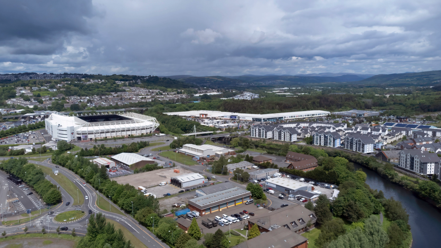 Aerial view of Swansea, showing a stadium, industrial area, residential blocks along a river, and distant hills under a cloudy sky.