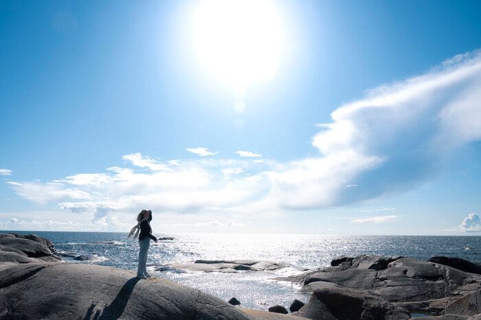 En person står på en klippefyldt kyst og kigger op på den klare sol over det funklende hav.