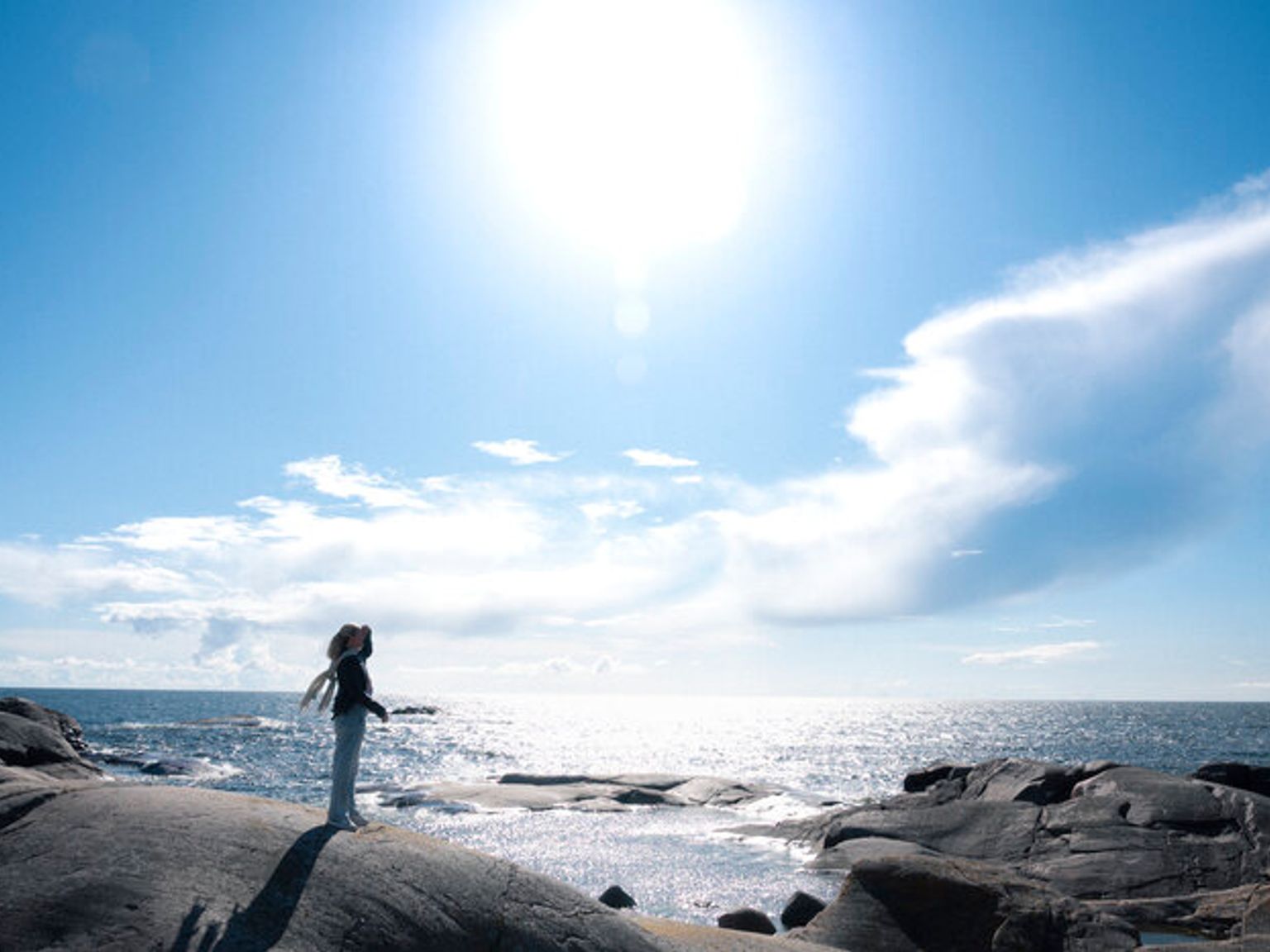 En person står på en klippefyldt kyst og kigger op på den klare sol over det funklende hav.