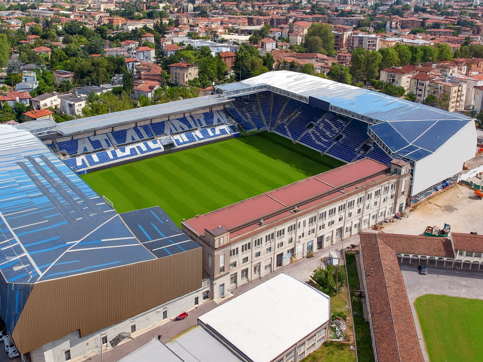 Ilmakuva modernista stadionista, jossa on vihreä kenttä ja siniset istuimet, jotka muodostavat sanan "ATALANTA", ympäröitynä kaupunkimaisemalla.