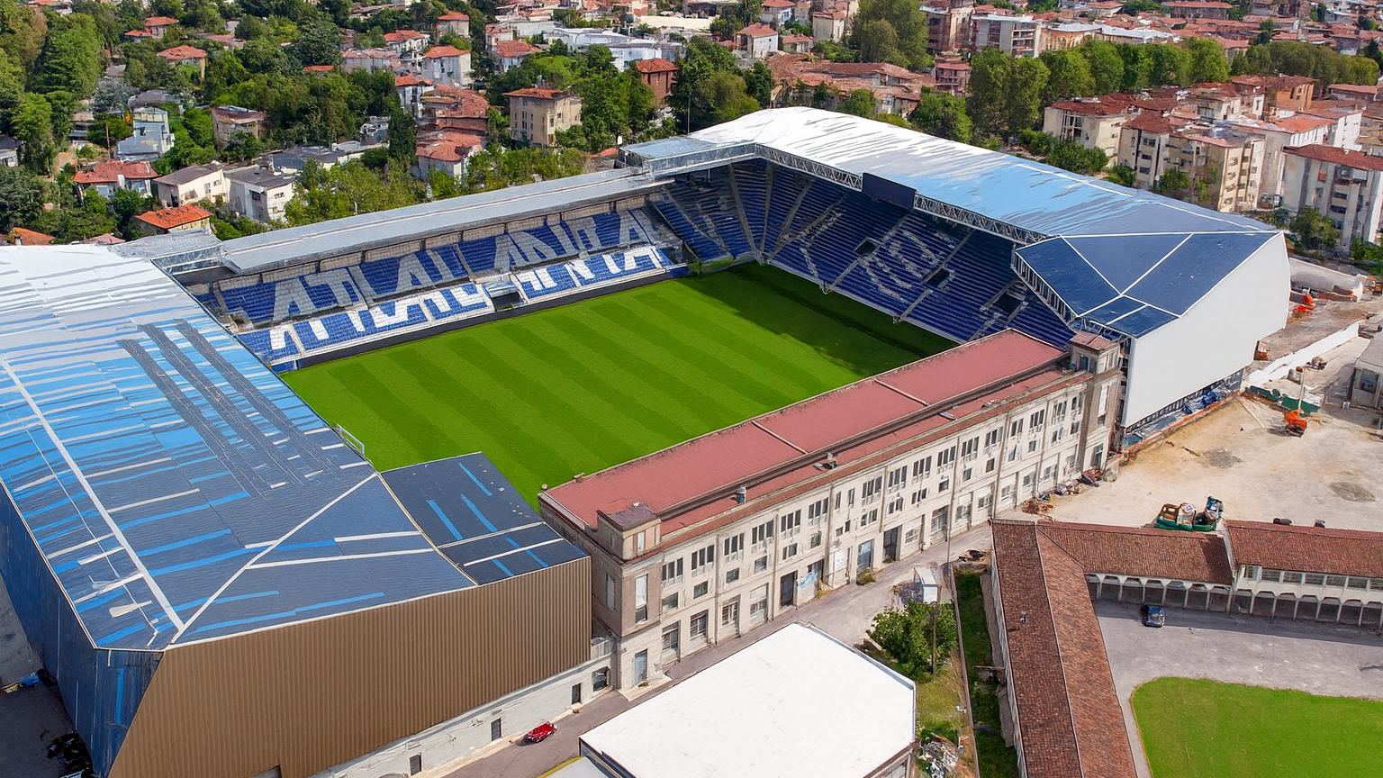 Aerial view of a modern stadium with a green pitch and blue seats spelling "ATALANTA," surrounded by an urban landscape.