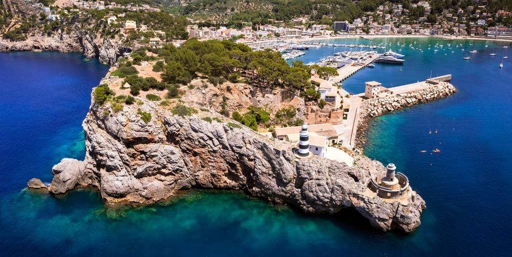 Luftfoto af en klippefyldt halvø med et stribet fyrtårn med udsigt over en marina og en kystby. Sóller, Mallorca