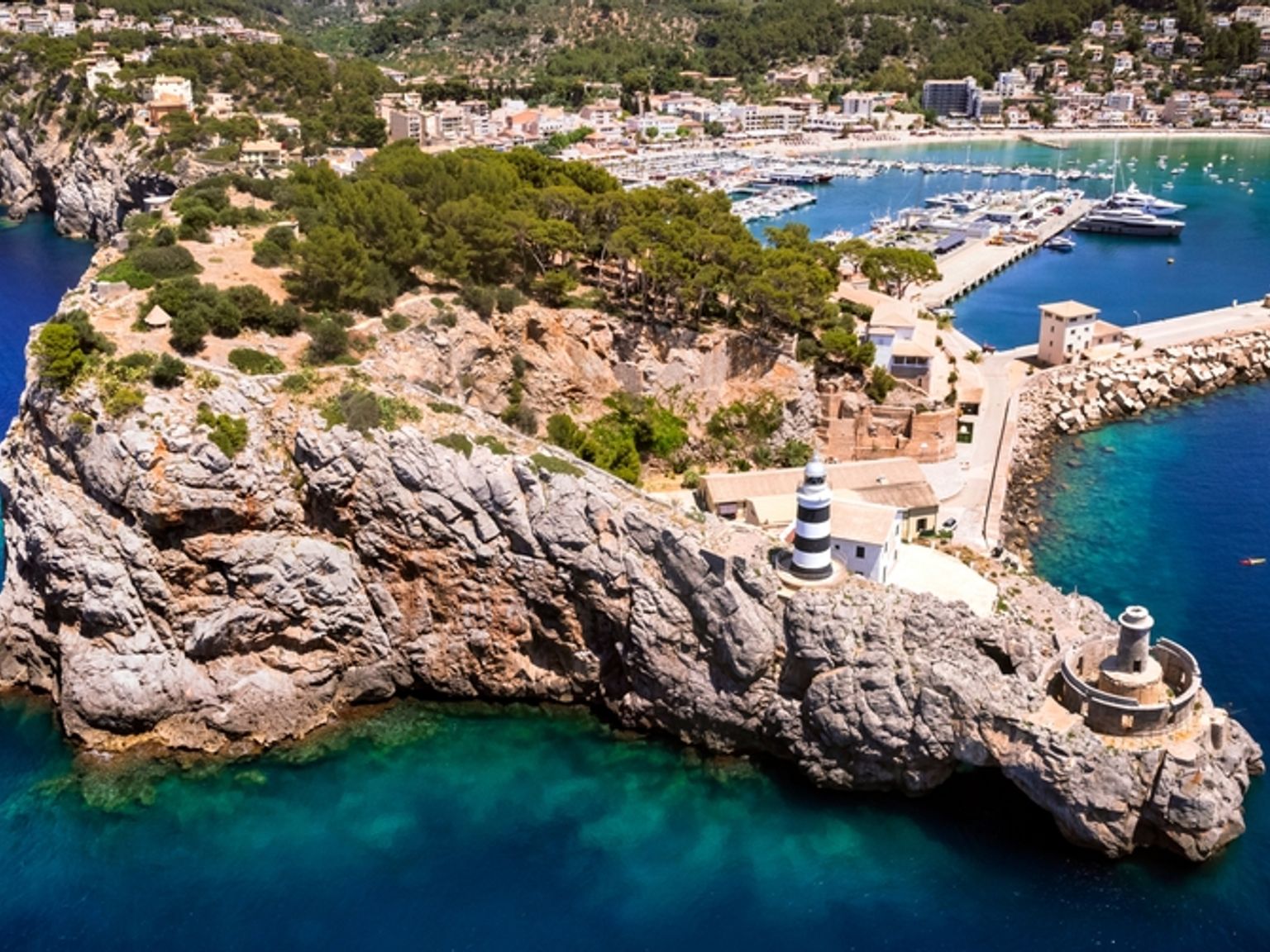 Flygvy över en stenig halvö med en randig fyr som blickar ut över en marina och en kuststad. Sóller, Mallorca