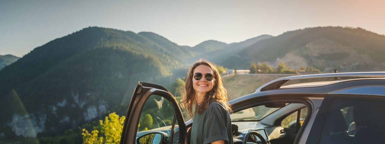 Smilende kvinne med solbriller ved siden av en åpen bil, fjell ved solnedgang.