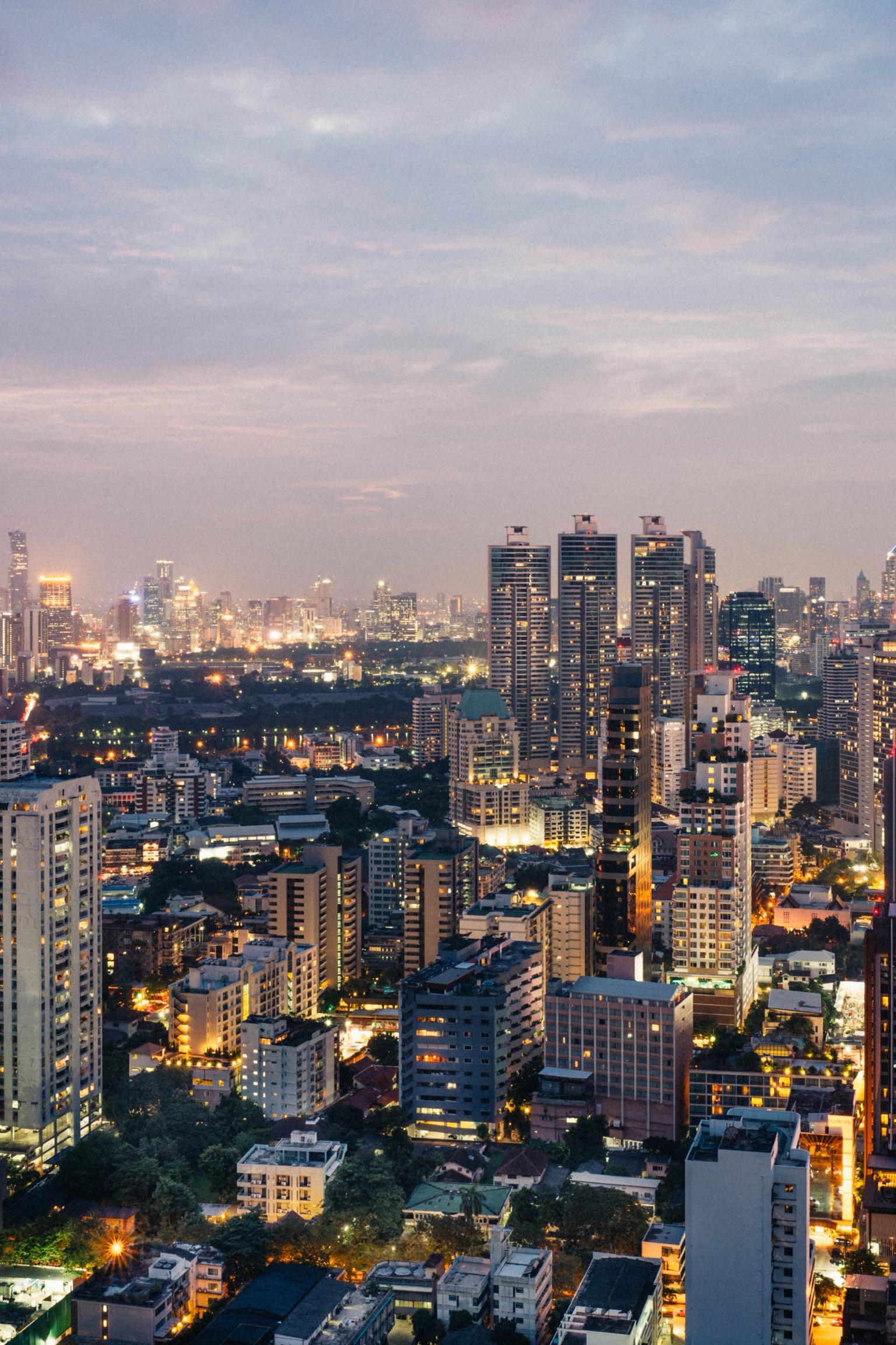 Bangkok ved skumring med adskillige oplyste bygninger under en skyet himmel.