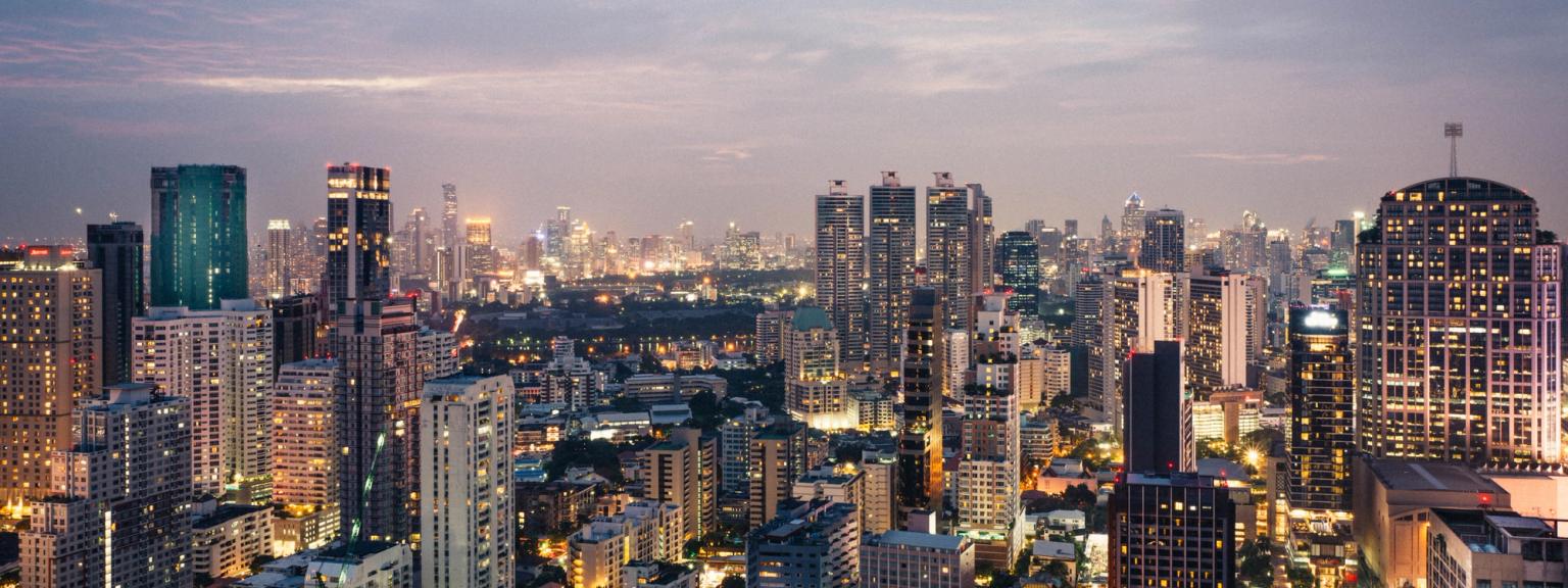 Bangkok ved skumring med adskillige oplyste bygninger under en skyet himmel.