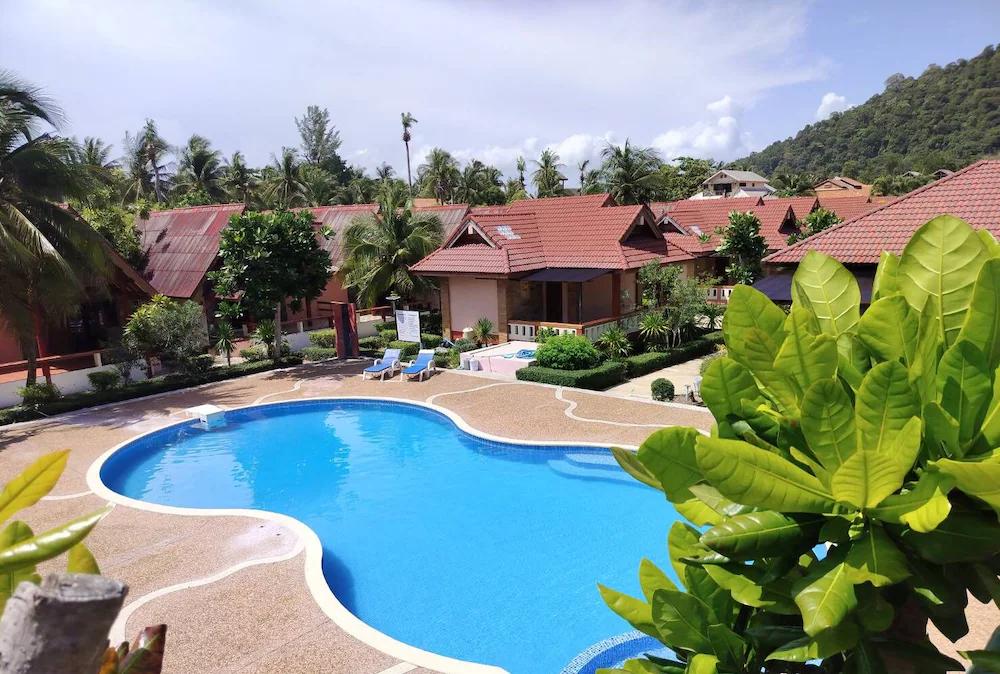 Tropiskt resort med en blå swimmingpool, bungalower med röda tak, palmer och ett skogsklätt berg.