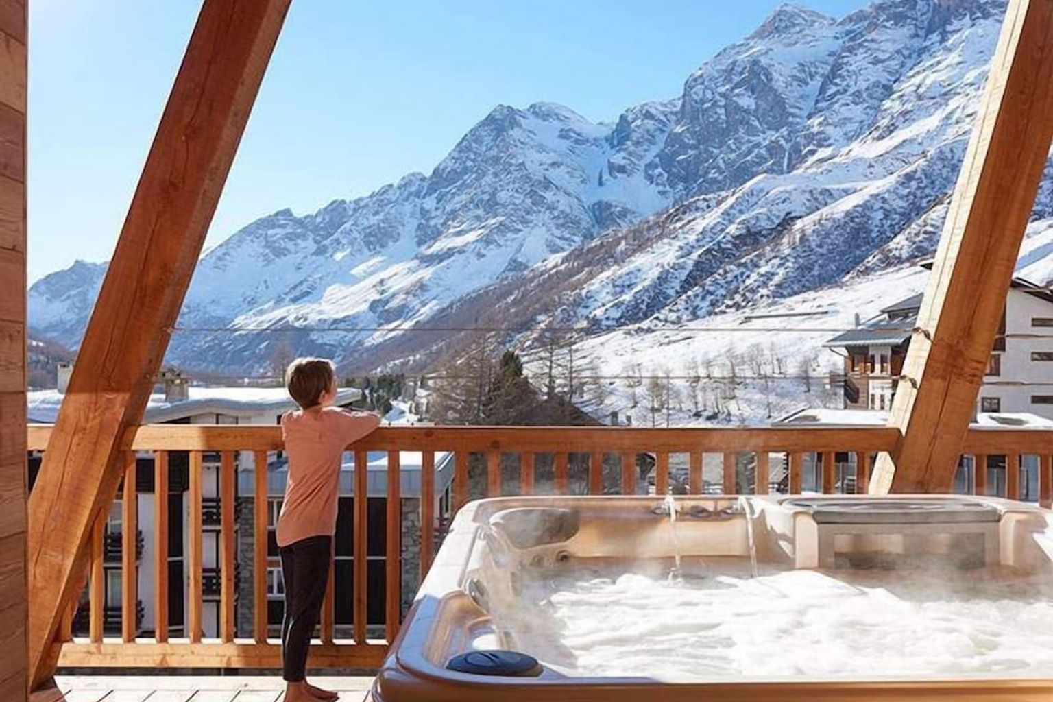 Et barn står på en trebalkong med et dampende boblebad og ser utover snødekte fjell.