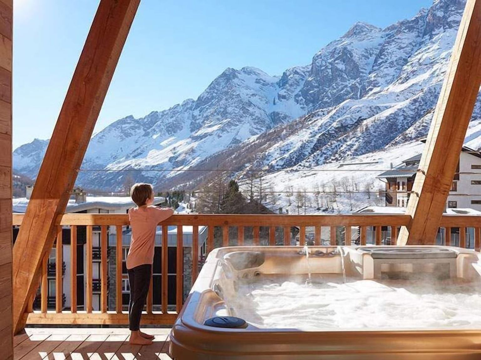 Et barn står på en trebalkong med et dampende boblebad og ser utover snødekte fjell.