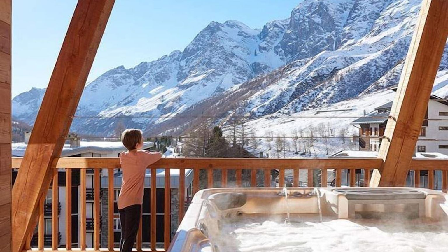 Et barn står på en trebalkong med et dampende boblebad og ser utover snødekte fjell.