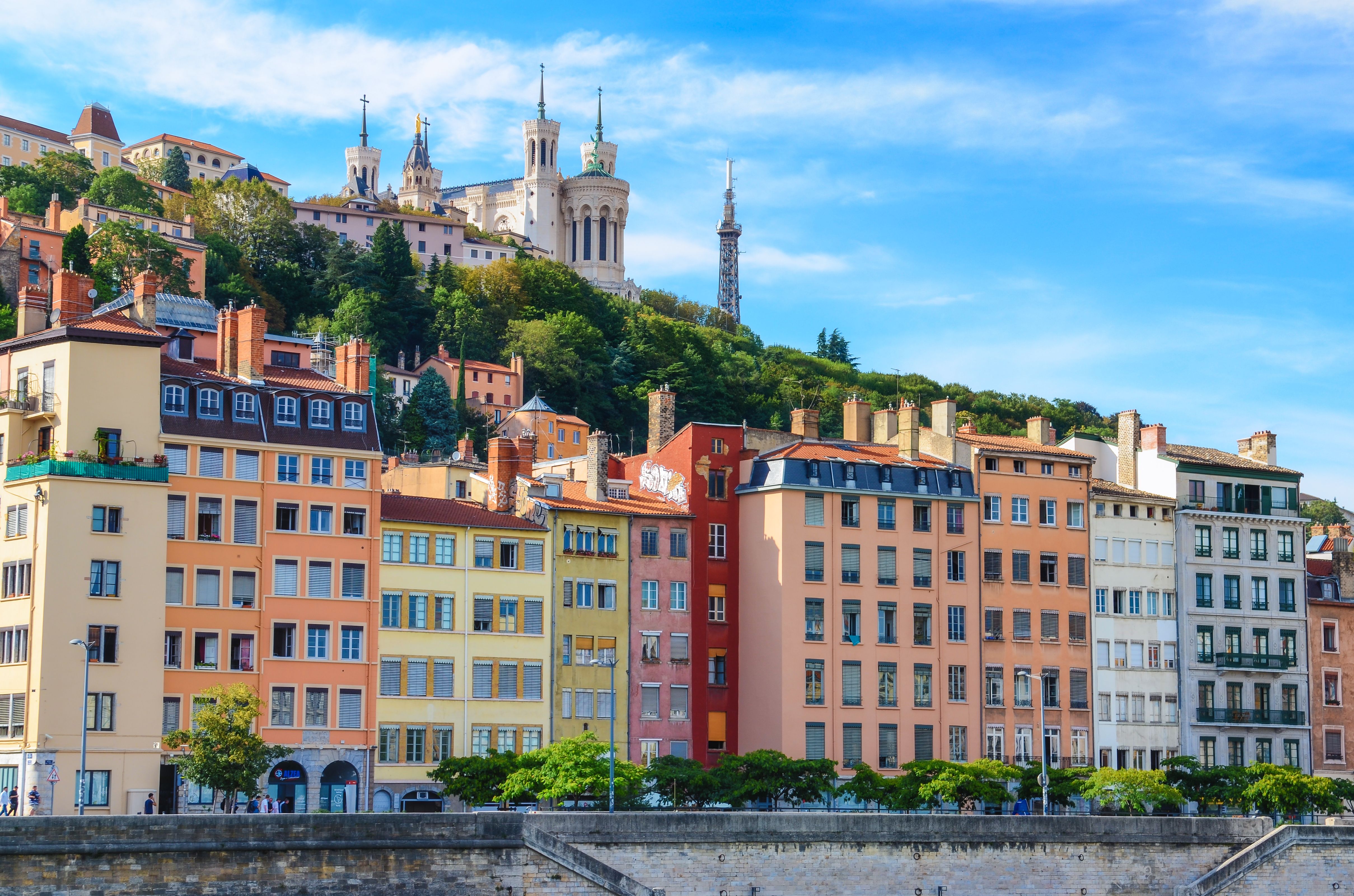 Färgglada byggnader längs en flod med Lyons Fourvièrebasilika på en kulle.