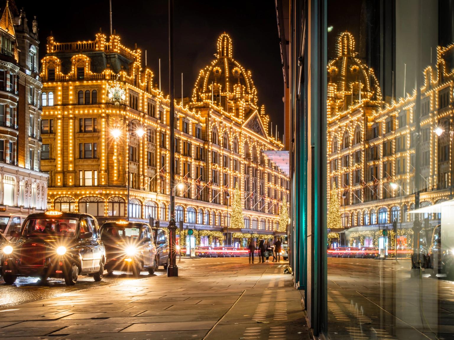 Harrods varuhus i London är lysande upplyst med gyllene ljus på natten, med svarta taxibilar och en fönsterreflektion.