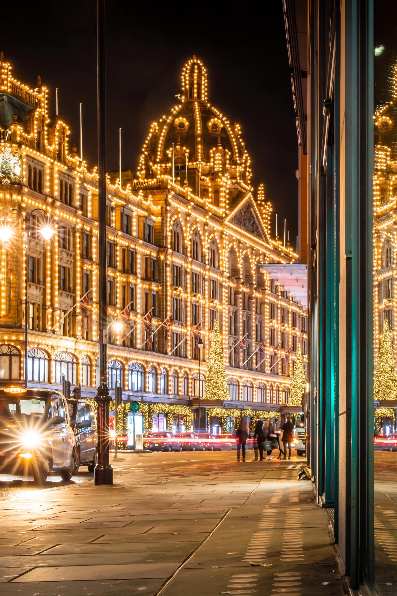 Harrods varuhus i London är lysande upplyst med gyllene ljus på natten, med svarta taxibilar och en fönsterreflektion.
