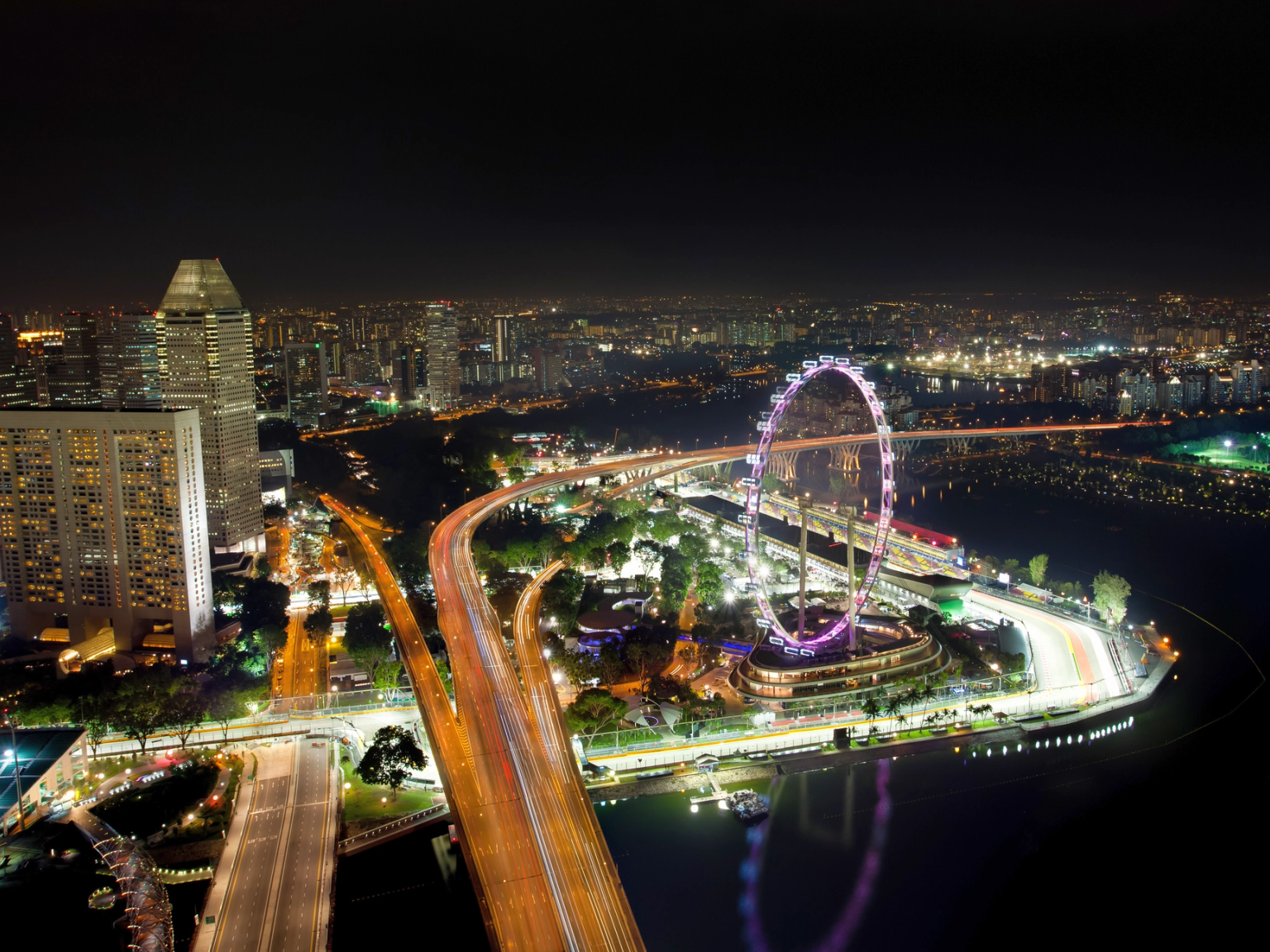 Éjszakai légi felvétel Szingapúr pezsgő városképéről, a kivilágított Singapore Flyer óriáskerékkel, fénycsíkos autópályákkal és a Marina-öböllel.