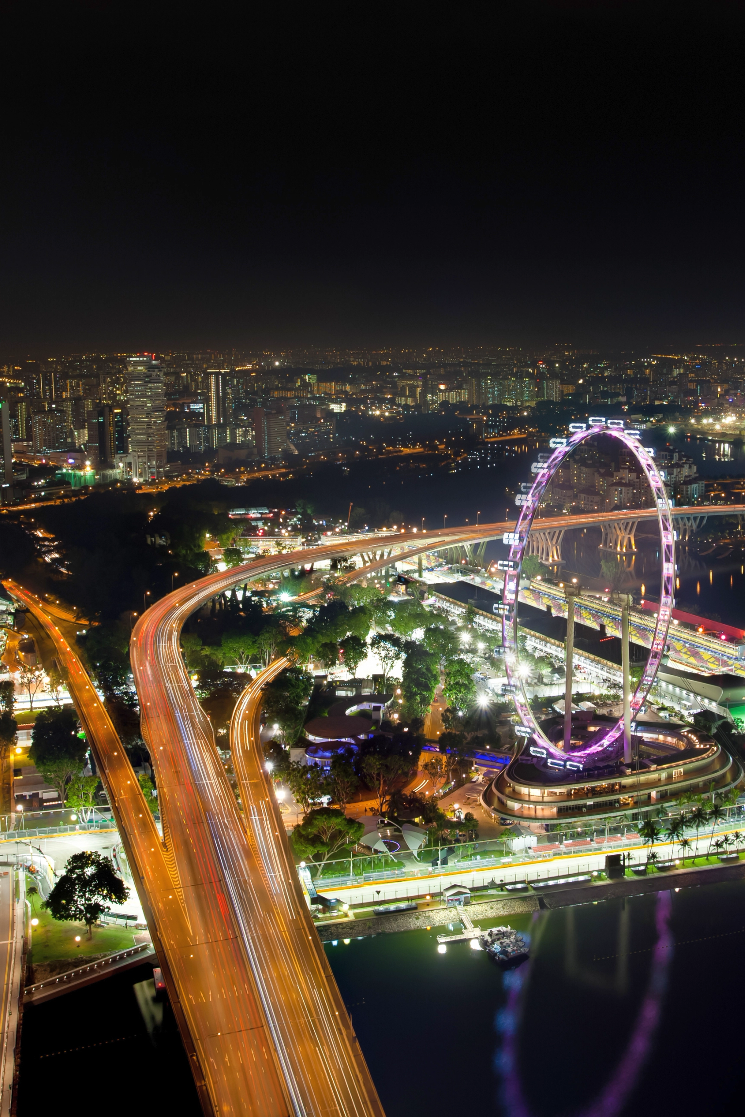Éjszakai légi felvétel Szingapúr pezsgő városképéről, a kivilágított Singapore Flyer óriáskerékkel, fénycsíkos autópályákkal és a Marina-öböllel.