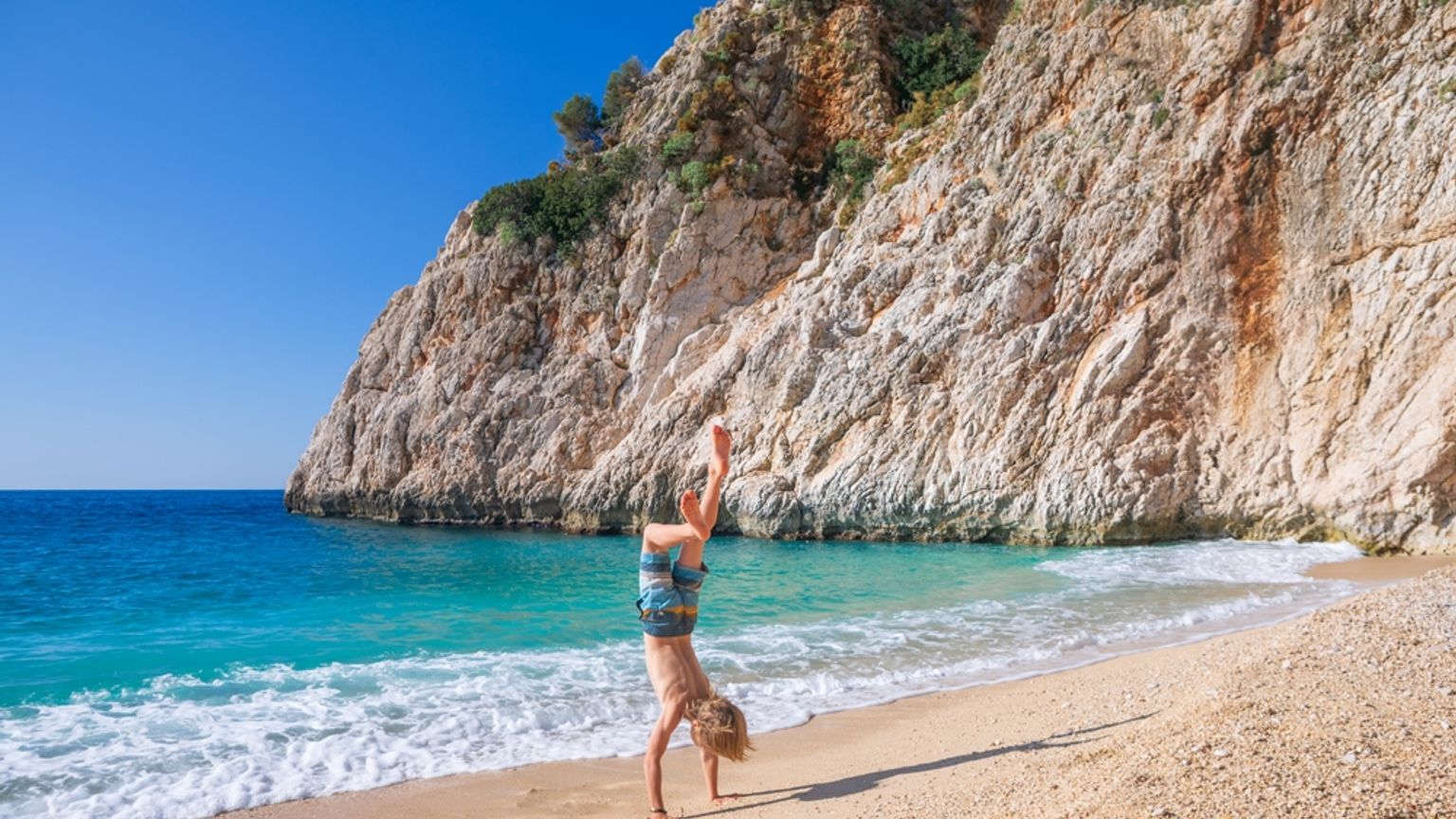 Et barn laver en håndstand på en sandstrand med turkis vand og en stor klippe. Antalya - Tyrkiet