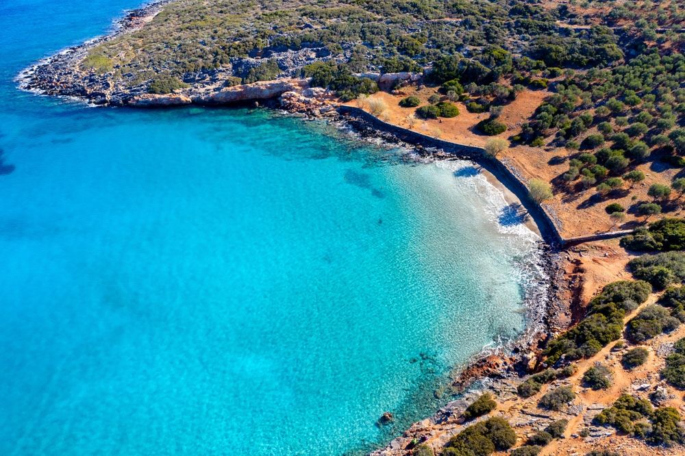 Luchtfoto van een afgelegen turkooizen baai met een zandstrand, ondersteund door een stenen muur en droog, met bomen bedekt land. Elounda, Griekenland