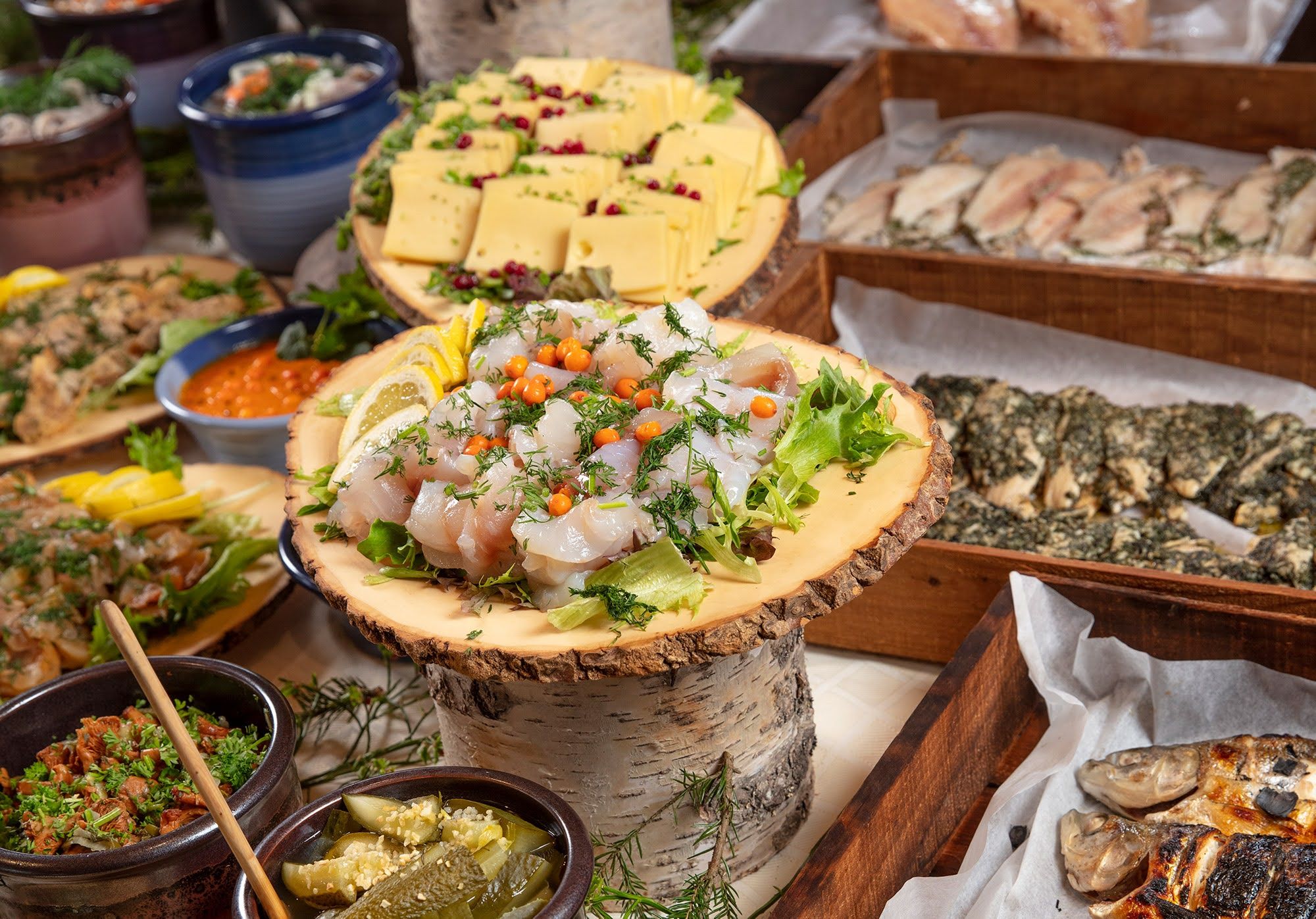 En rustik buffet med marineret fisk, skåret ost, grillet fisk og forskellige andre retter på træplanker og i skåle.