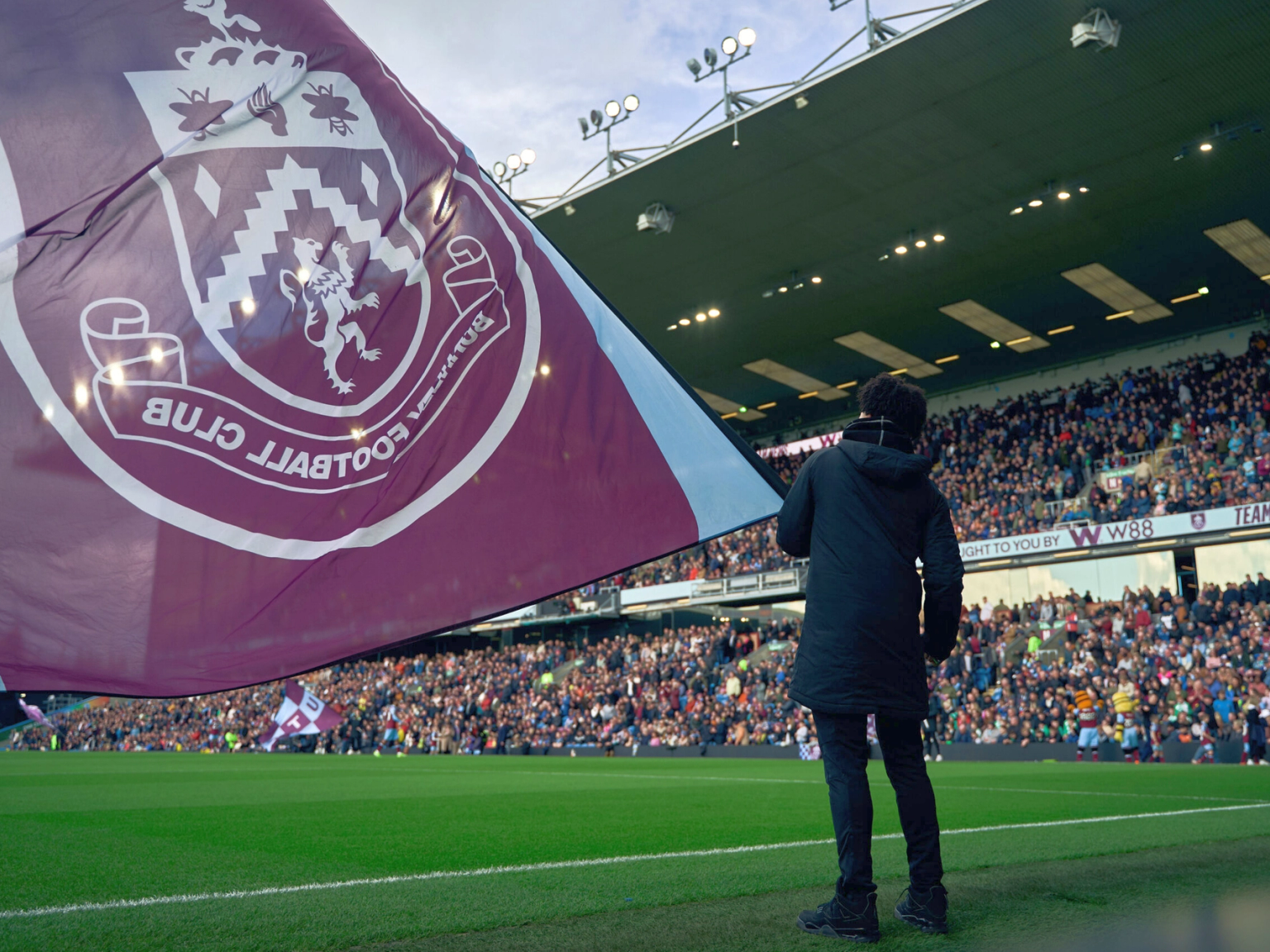 Uma grande bandeira do Burnley F.C. desdobrada sobre um campo de futebol, parcialmente obscurecendo uma pessoa de costas para a câmera, voltada para as arquibancadas lotadas do estádio.