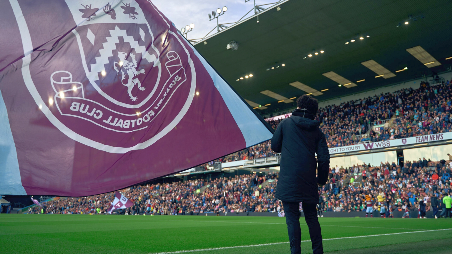 Un grand drapeau du Burnley F.C. déployé sur un terrain de football, cachant partiellement une personne de dos à la caméra, face aux tribunes bondées du stade.