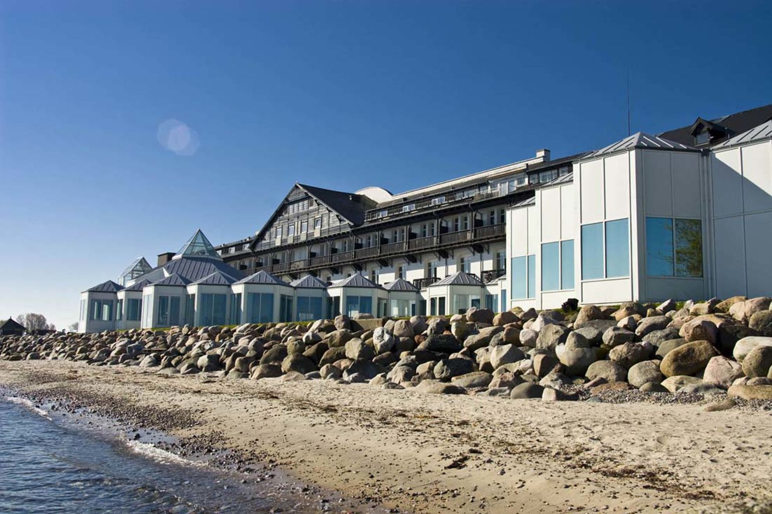 En stor resortbyggnad på en sandstrand med en stenbarriär vid vattnet. Marienlyst Strandhotel