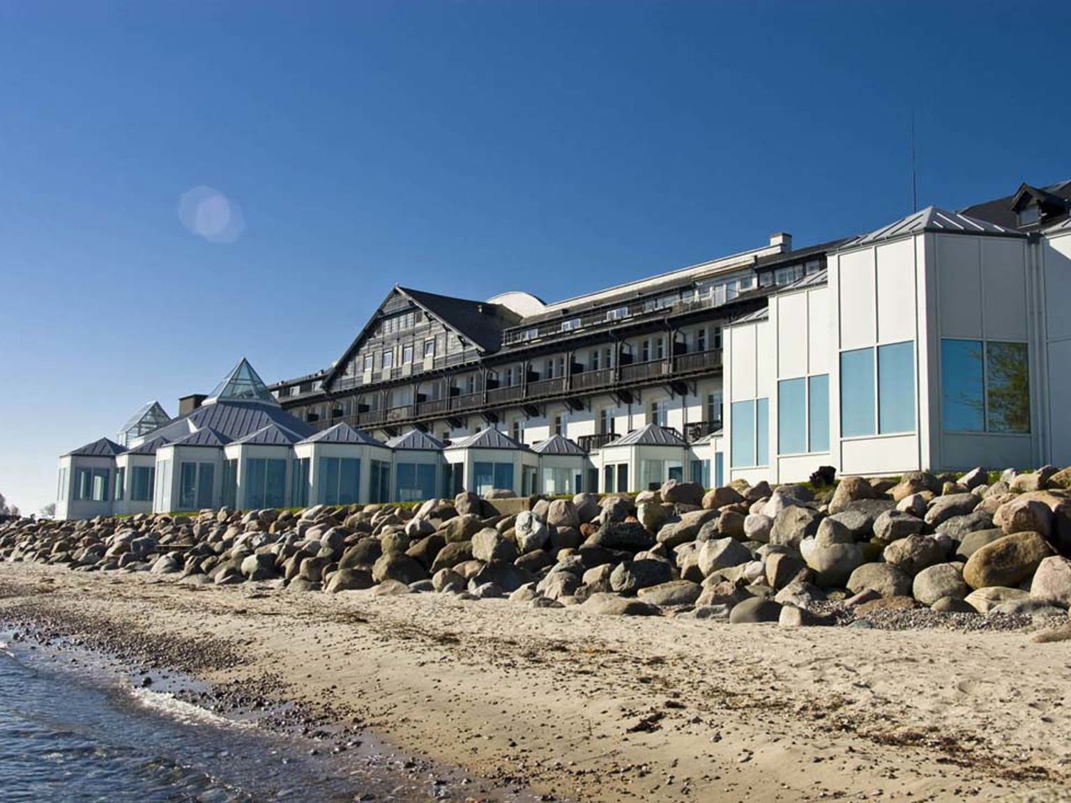 En stor resortbyggnad på en sandstrand med en stenbarriär vid vattnet. Marienlyst Strandhotel