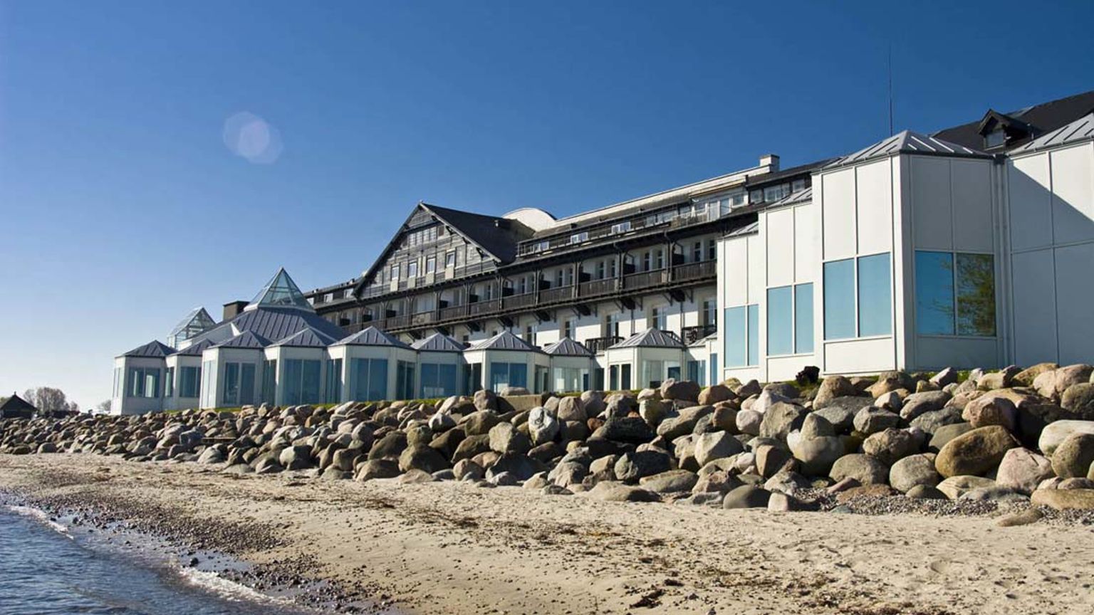 En stor resortbyggnad på en sandstrand med en stenbarriär vid vattnet. Marienlyst Strandhotel