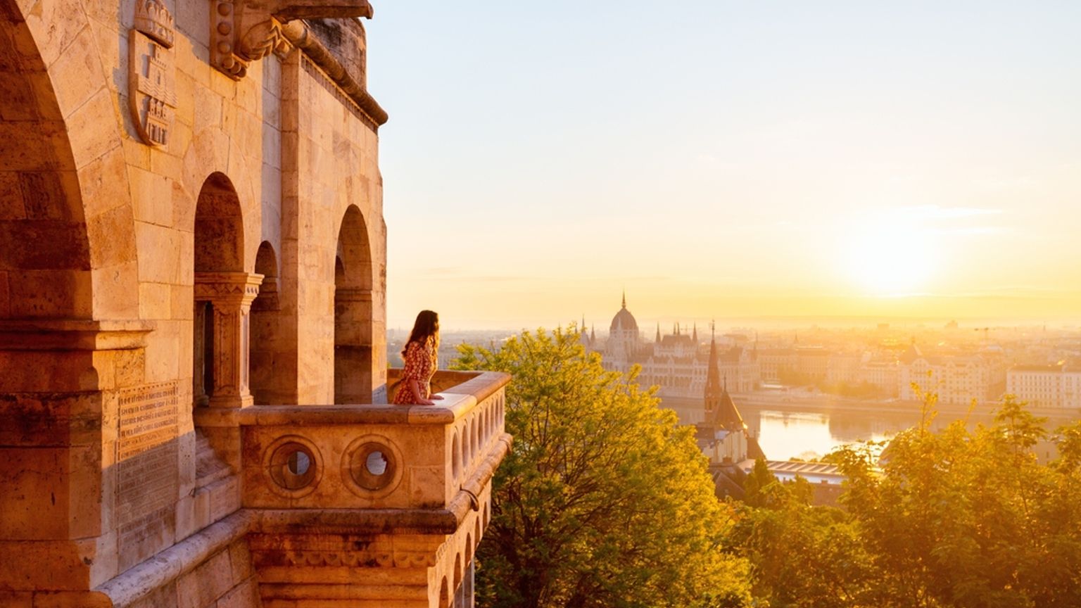 Kvinna på en stenbalkong som ser soluppgången över Budapest, med parlamentsbyggnaden och floden Donau synliga.