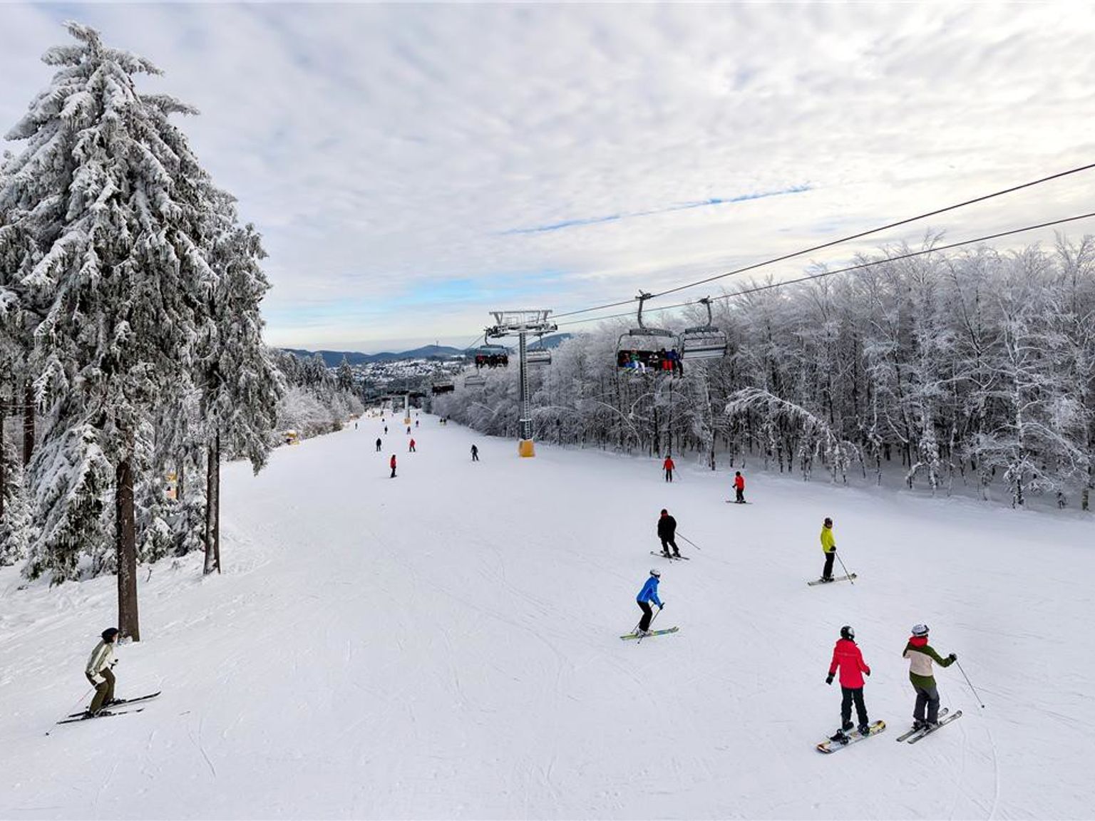 Skiërs en snowboarders dalen een besneeuwde berghelling af, geflankeerd door met rijp bedekte bomen, met een skilift zichtbaar boven hen. Center Parcs Park Hochsauerland