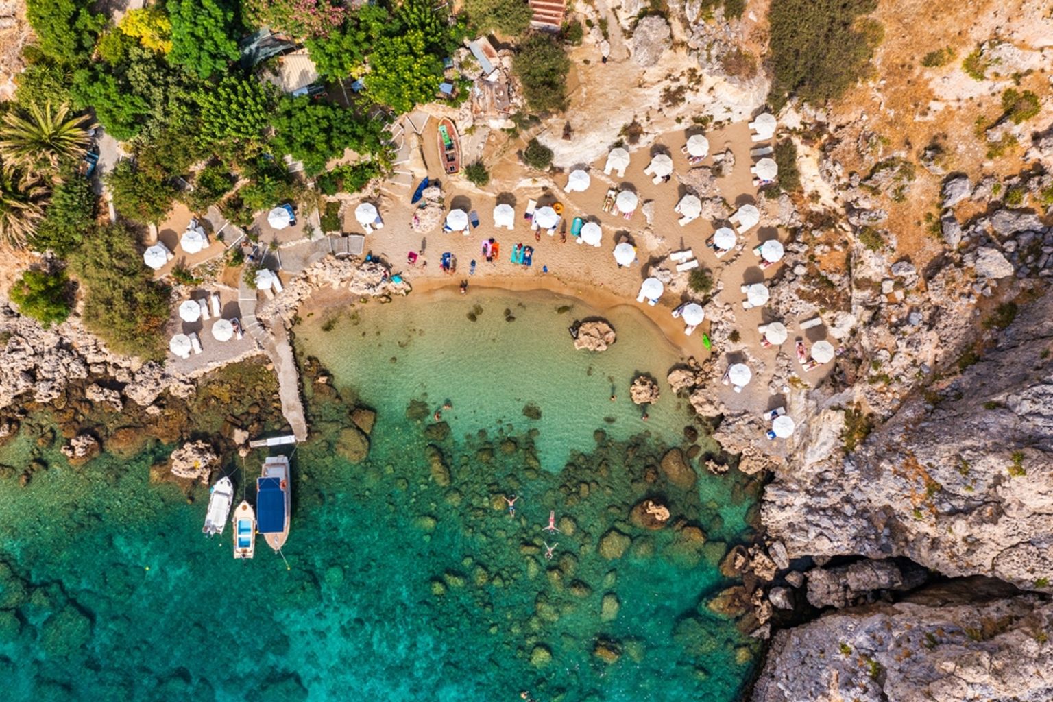 Flyfoto av en avsides strand med hvite parasoller, klart turkis vann og båter. Rhodos, Hellas