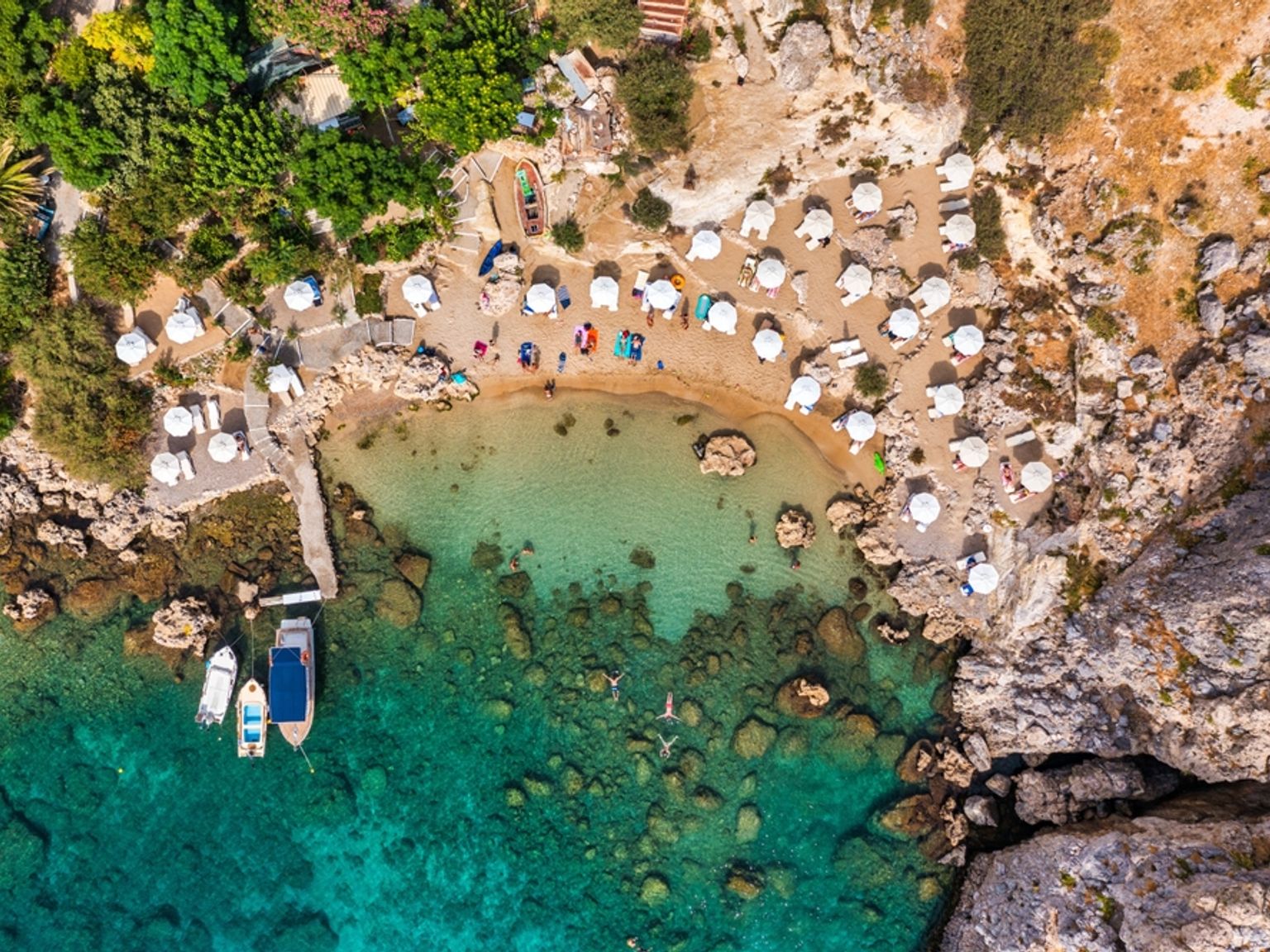 Flyfoto av en avsides strand med hvite parasoller, klart turkis vann og båter. Rhodos, Hellas