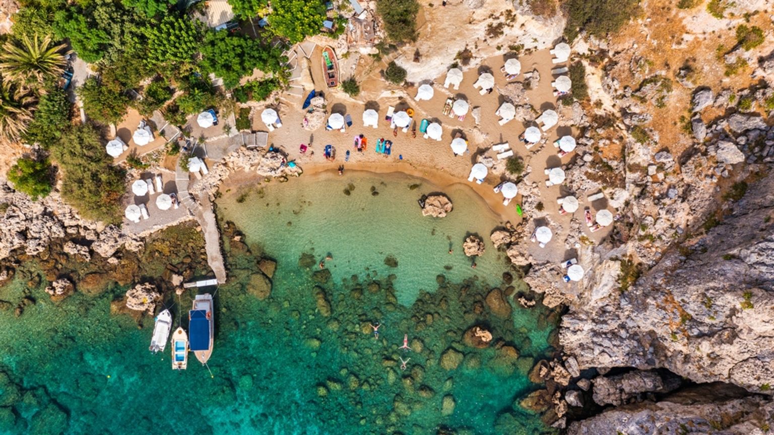 Flyfoto av en avsides strand med hvite parasoller, klart turkis vann og båter. Rhodos, Hellas